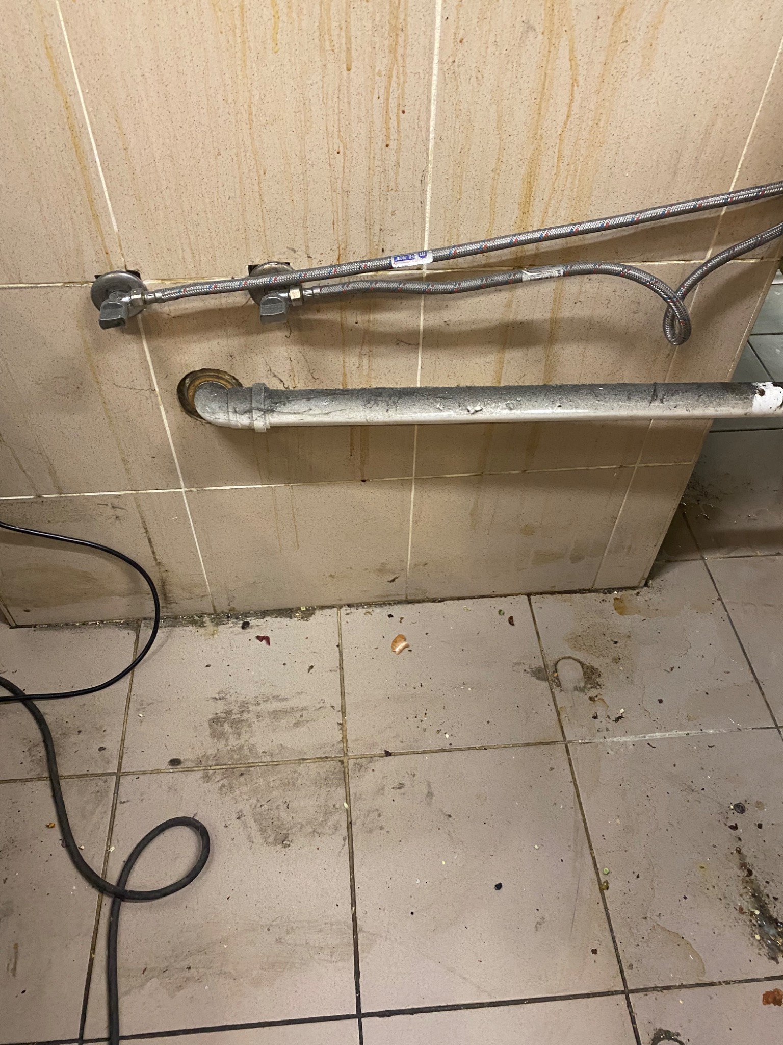 Brudna podłoga i ściany w łazience przed sprzątaniem, widoczne rury i wężyki doprowadzające wodę, ślady po zaciekach i zabrudzeniach na płytkach.