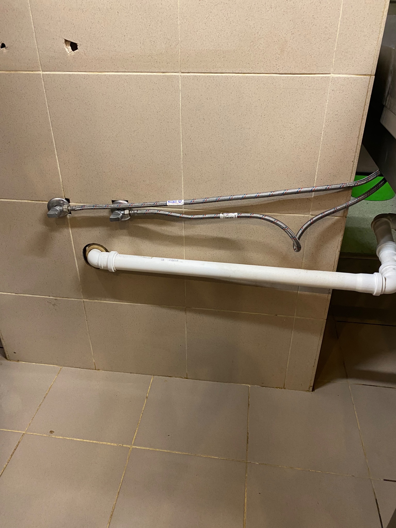 Fragment ściany z beżowych płytek z widocznymi otworami, wystającymi rurami i przewodami instalacyjnymi, sugerujący stan przed lub w trakcie prac remontowych.