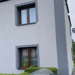 Dwukondygnacyjny dom z jasną elewacją i szarymi akcentami wokół okien i na narożnikach, widoczna skrzynka pocztowa i żywopłot w ogrodzie.