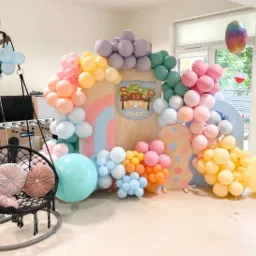 Dekoracja balonowa w pastelowych kolorach, tworząca łuk wokół drewnianej ścianki z herbem Zgierza, uzupełniona wiszącym fotelem z poduszkami i dodatkowymi balonami, tworząca scenografię imprezową.