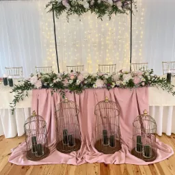 Dekoracja stołu weselnego z kwiatami, świecami w szklanych lampionach w kształcie klatek i podświetlanym tłem z białej tkaniny.