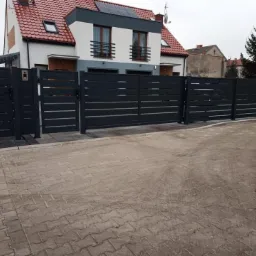 Nowoczesne, poziome ogrodzenie metalowe w antracytowym kolorze, z przesuwną bramą wjazdową i furtką, na tle domu z czerwoną dachówką i szarą elewacją.