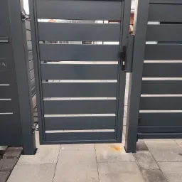 Nowoczesna, grafitowa brama wjazdowa z poziomymi panelami, z widocznym zamkiem i klamką, osadzona na kostce brukowej w odcieniach szarości.