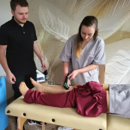 Kobieta w szarym uniformie aplikuje urządzenie do terapii ultradźwiękowej na nodze pacjenta leżącego na kozetce; asystuje mężczyzna w czarnej koszulce polo, w tle ściana z motywem piór.