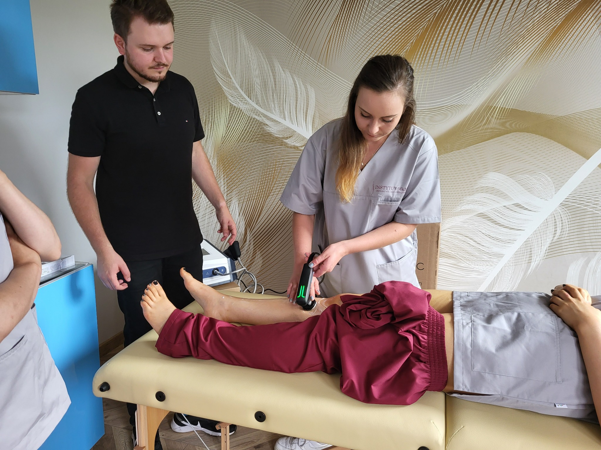 Kobieta w szarym uniformie aplikuje urządzenie do terapii ultradźwiękowej na nodze pacjenta leżącego na kozetce; asystuje mężczyzna w czarnej koszulce polo, w tle ściana z motywem piór.