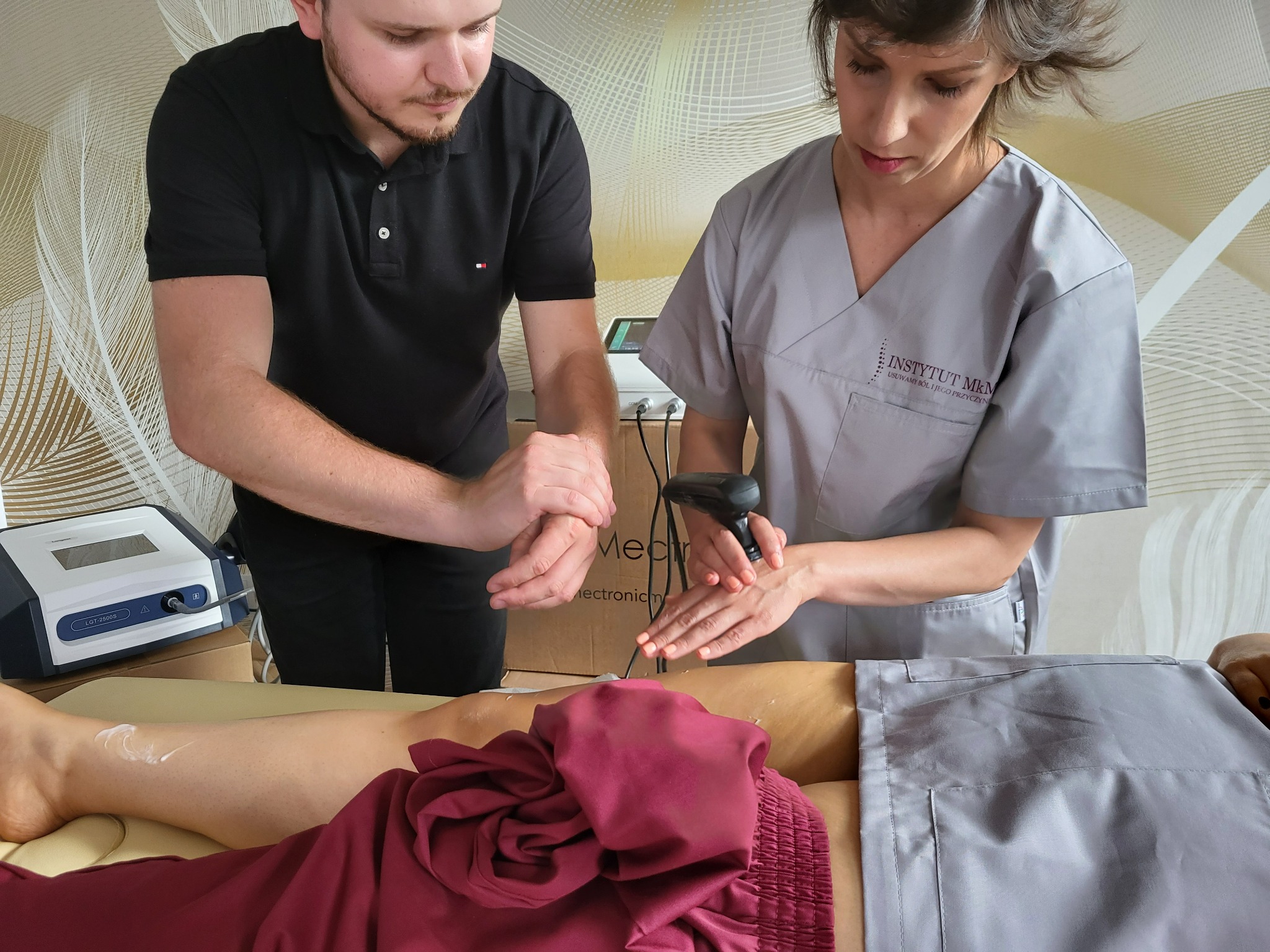 Dwoje specjalistów w trakcie zabiegu na nodze pacjenta, wykorzystujących urządzenie do elektroterapii. Widoczny krem na skórze i logo 'Instytut Mkm' na uniformie kobiety.