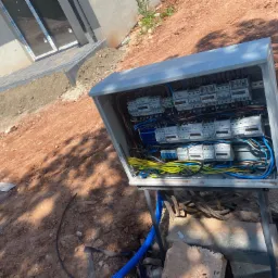 Otwarta skrzynka elektryczna z podłączonymi przewodami i licznikami na zewnątrz budynku w trakcie montażu, widoczne niebieskie rury karbowane i ziemiste podłoże.