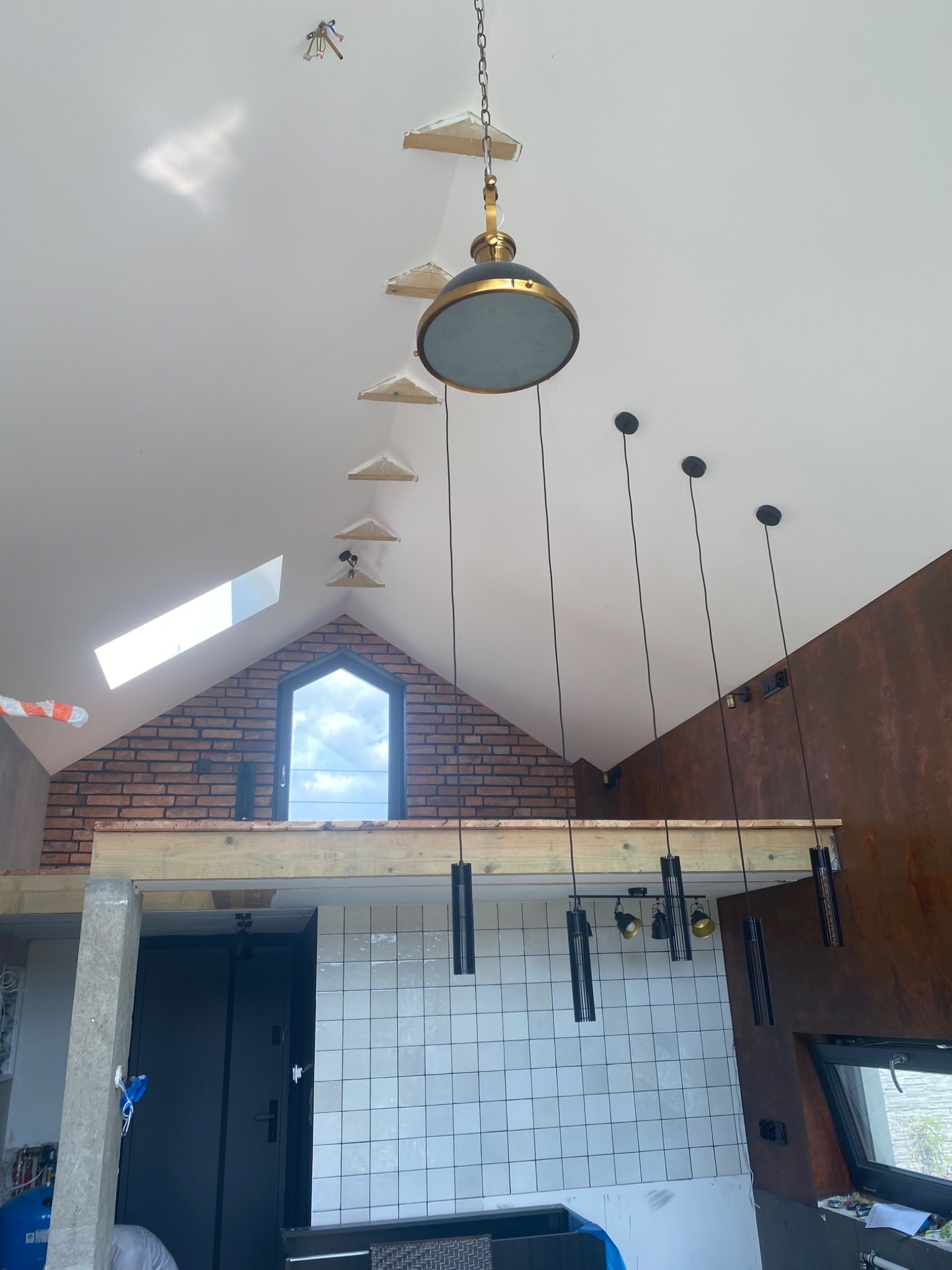Wnętrze domu w trakcie wykańczania z widocznym oświetleniem: centralnie umieszczona lampa wisząca z elementami w kolorze złota, rząd lamp wiszących na czarnych kablach, fragment ściany z cegły...