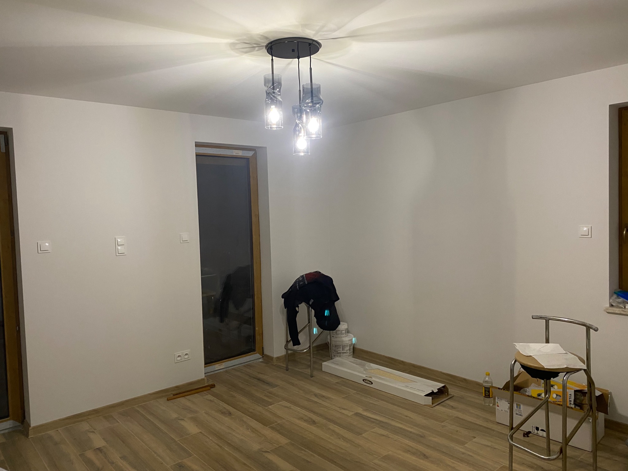 Nowoczesna lampa wisząca z trzema szklanymi kloszami w surowym wnętrzu z drewnianą podłogą, widoczne puszki po farbie i krzesło z kartonami.