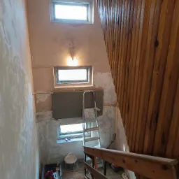 Klatka schodowa w trakcie remontu: ściany częściowo pokryte gładzią, widoczne okna, grzejnik, drabina, drewniana boazeria i narzędzia w kuwecie.