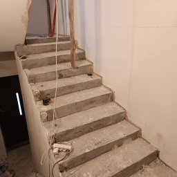 Renovation Usługi Remontowo-Budowlane Krystian Pajek - Surowe betonowe schody w trakcie remontu z widocznymi śladami prac murarskich i elektrycznych, wsparte drewnianymi belkami, z widocznymi narzędziami i przedłużaczem.