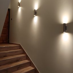 Renovation Usługi Remontowo-Budowlane Krystian Pajek - Klatka schodowa z drewnopodobnymi schodami, oświetlona trzema cylindrycznymi kinkietami emitującymi światło w górę i w dół, ściana pomalowana na jasny beż.