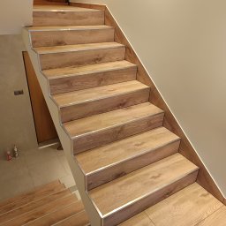 Renovation Usługi Remontowo-Budowlane Krystian Pajek - Schody wykończone panelami imitującymi drewno, z aluminiowymi listwami na krawędziach stopni, oświetlenie LED wzdłuż ściany, widok z dołu na górę.