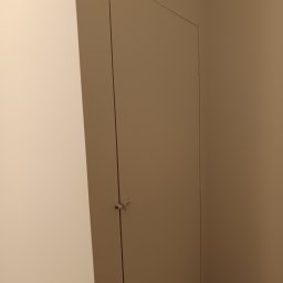 Renovation Usługi Remontowo-Budowlane Krystian Pajek - Ukryte drzwi do schowka na poddaszu z zamkiem baskwilowym, wtopione w ścianę pomalowaną na jednolity kolor.