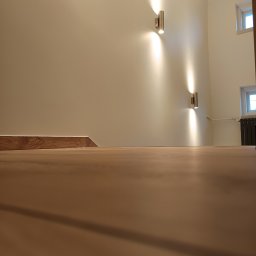 Renovation Usługi Remontowo-Budowlane Krystian Pajek - Widok z poziomu podłogi na świeżo pomalowaną ścianę z dwoma nowoczesnymi, metalowymi kinkietami, drewnianą listwą przypodłogową i grzejnikiem pod oknem.