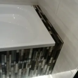 Obudowa wanny wykończona mozaiką w odcieniach szarości i brązu z podświetleniem LED, ściany wyłożone płytkami ceramicznymi w jasnym odcieniu.