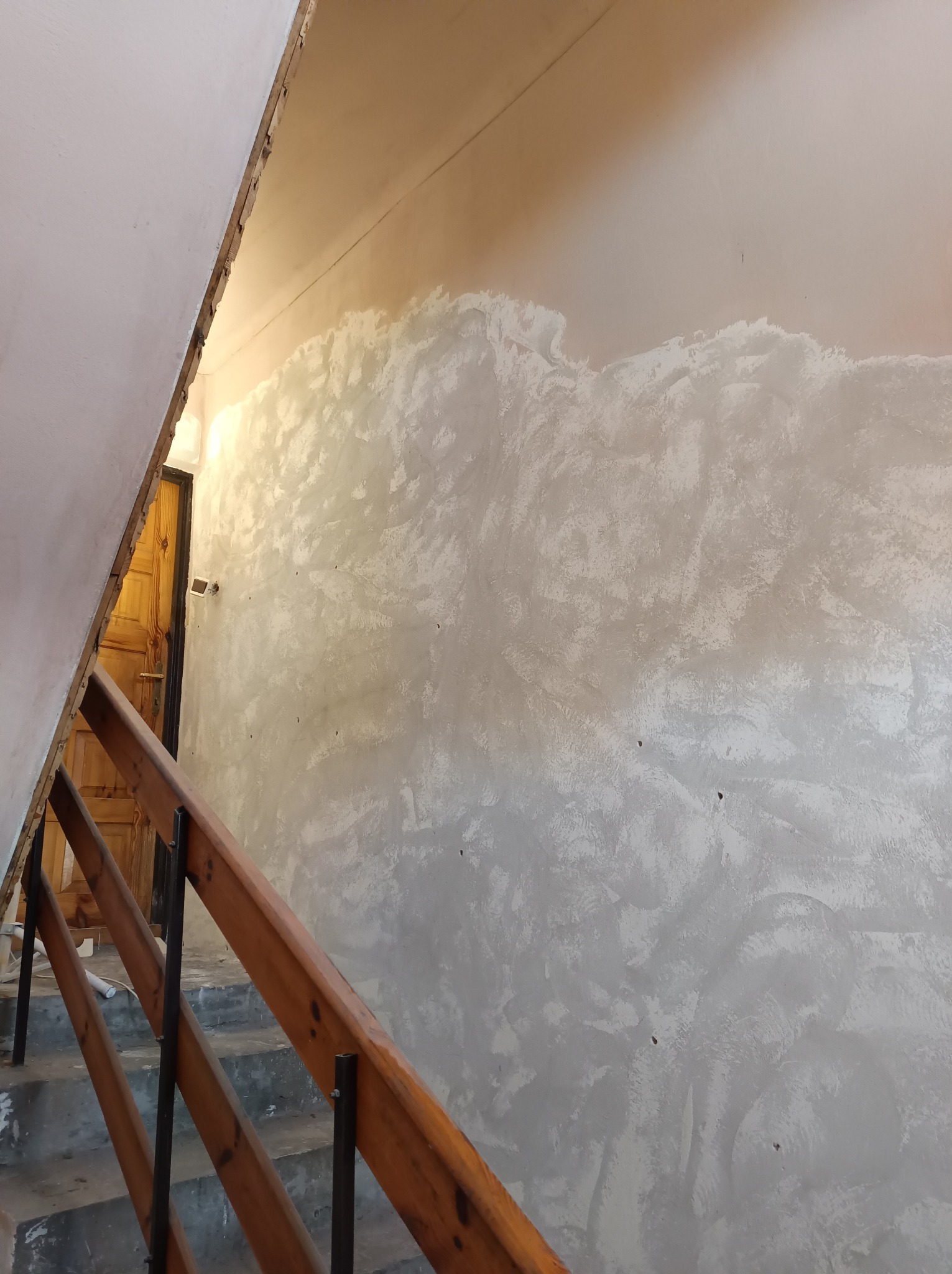 Ściana w trakcie nakładania gładzi, widoczne nierówności i faktura, schody z drewnianą barierką w tle, przygotowanie do malowania.