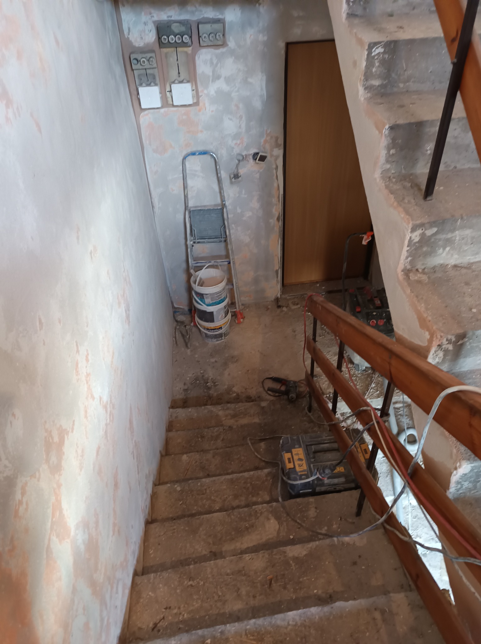 Klatka schodowa w trakcie remontu: ściany z widocznymi śladami szpachlowania, drabina, wiadro z farbą, skrzynka narzędziowa i kable elektryczne.