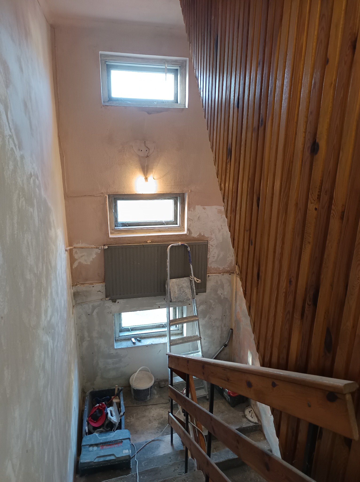 Klatka schodowa w trakcie remontu: ściany częściowo pokryte gładzią, widoczne okna, grzejnik, drabina, drewniana boazeria i narzędzia w kuwecie.