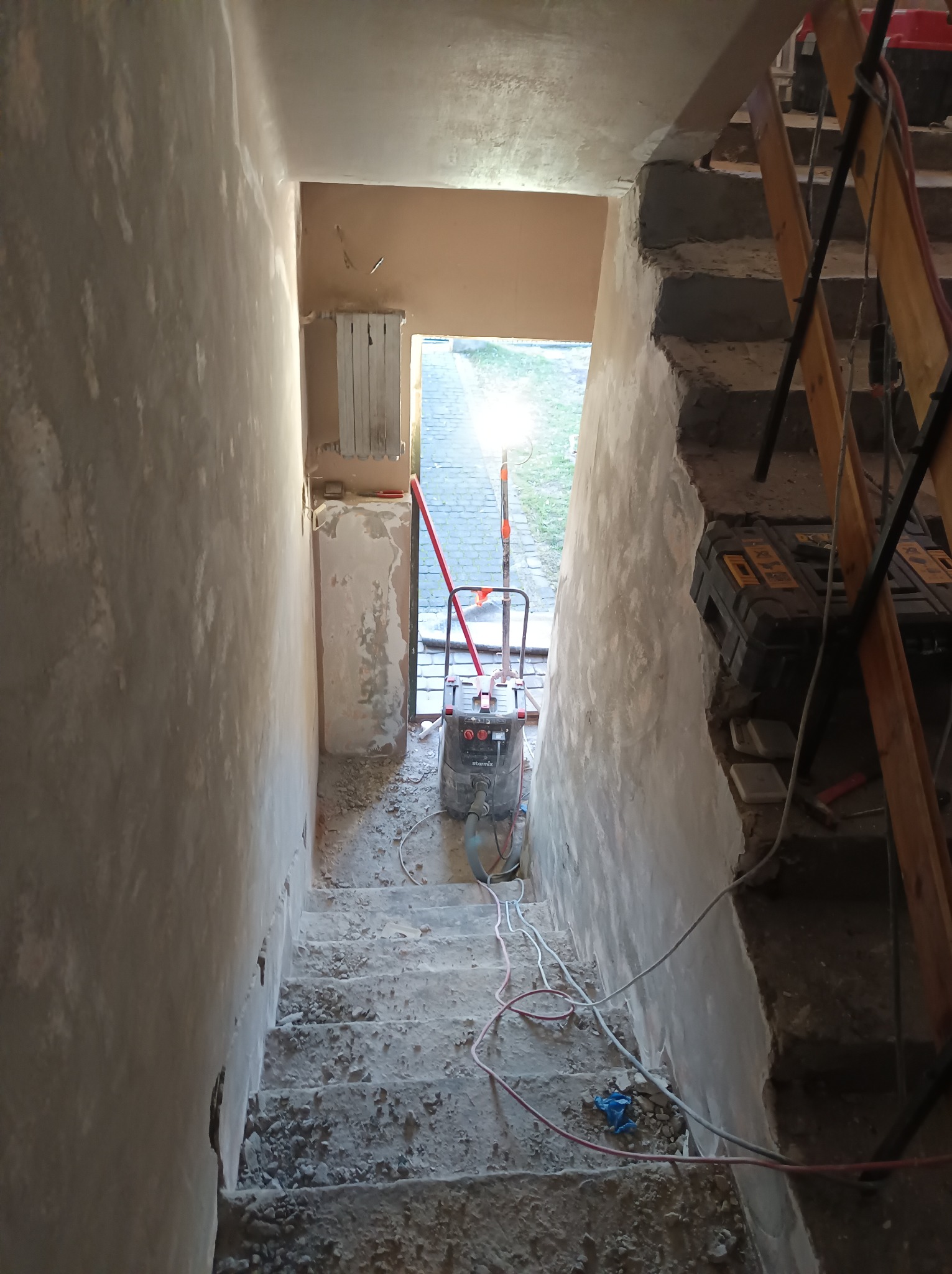 Klatka schodowa w trakcie gruntownego remontu: ściany w surowej gładzi, schody pokryte gruzem, widoczny agregat tynkarski, kable i narzędzia.