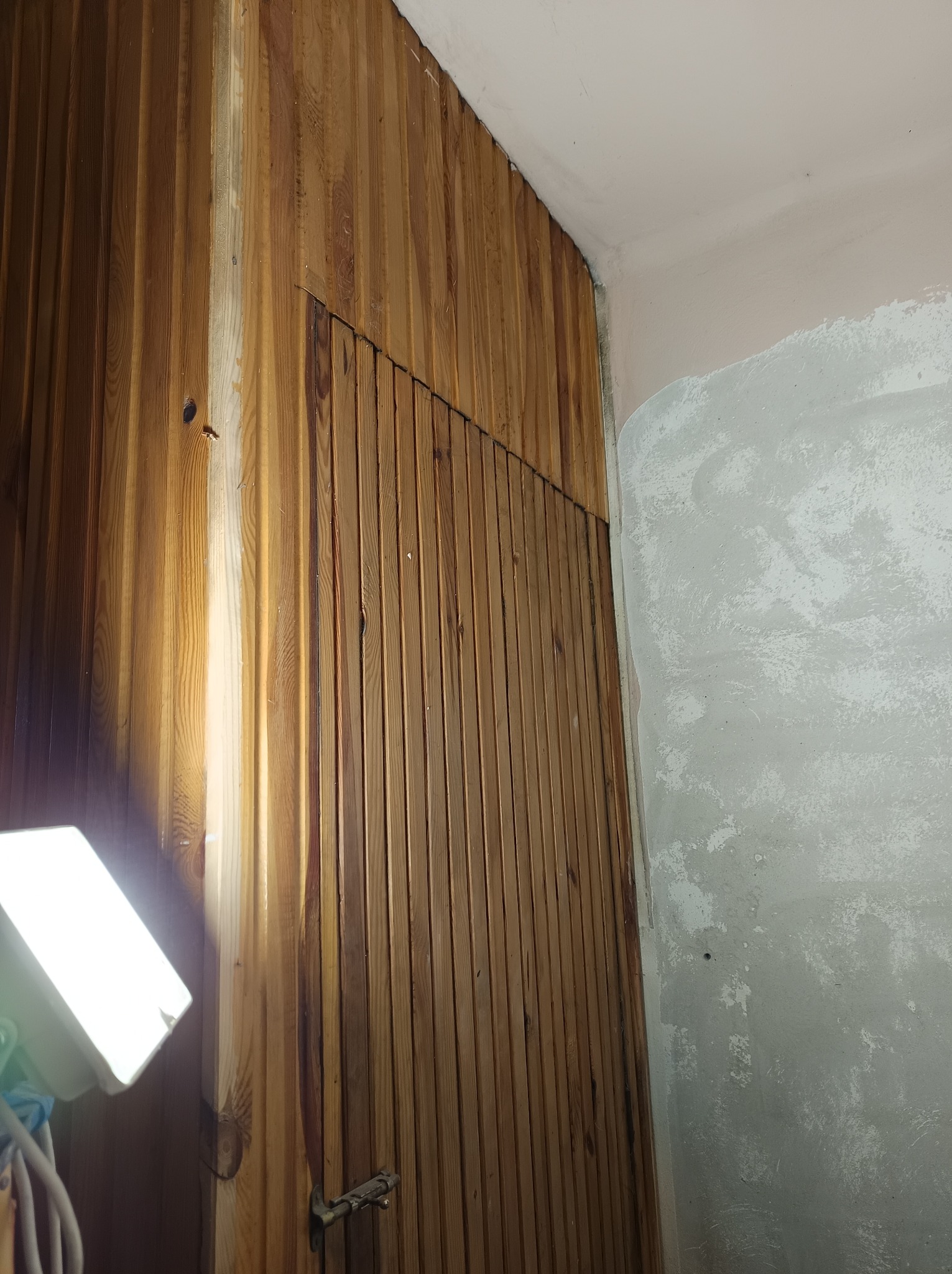Fragment ściany z boazerią w trakcie renowacji, widoczne ślady po starej powłoce i świeżo położone gładzie na sąsiedniej ścianie, oświetlane lampą roboczą.