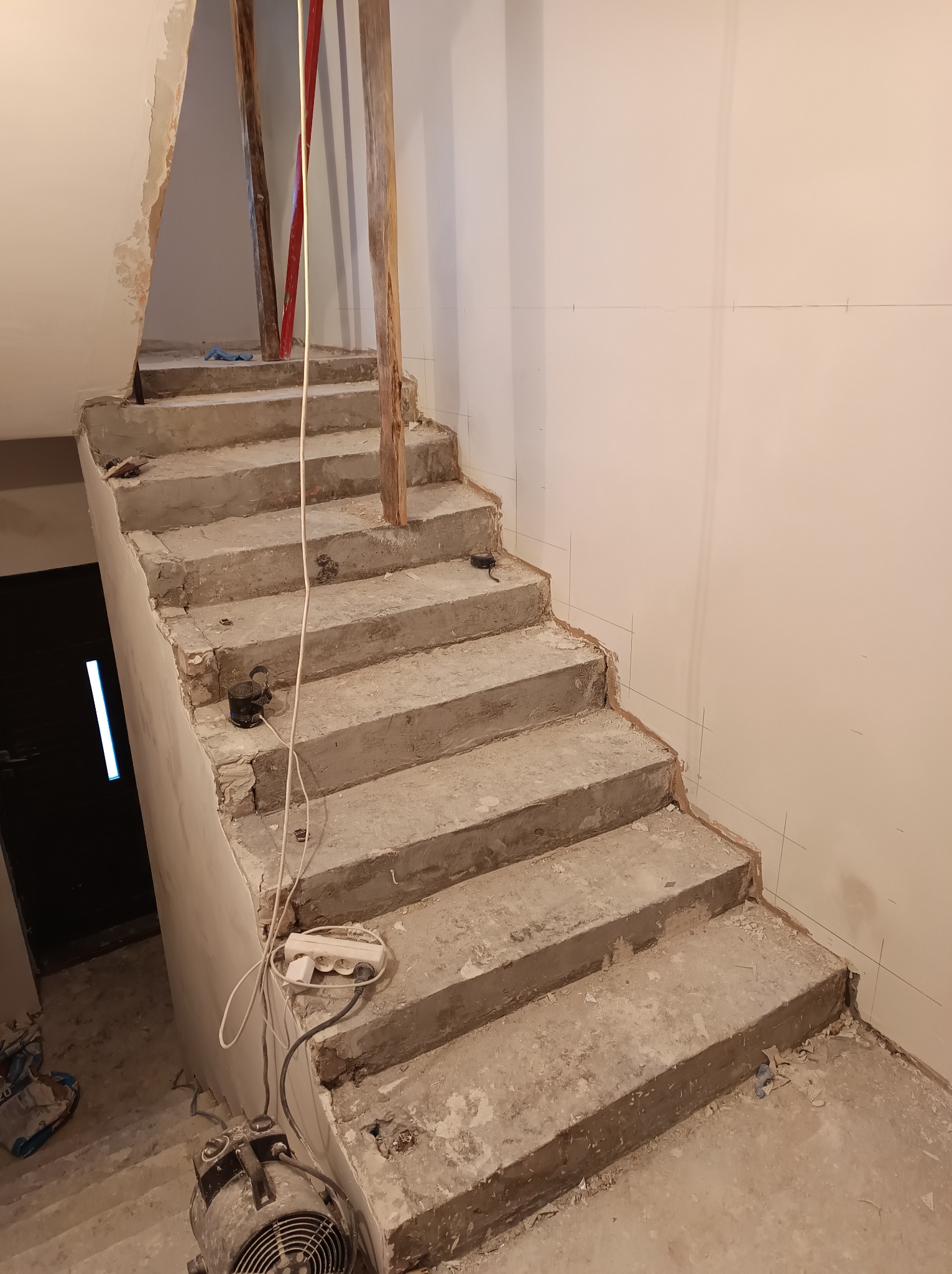 Surowe betonowe schody w trakcie remontu z widocznymi śladami prac murarskich i elektrycznych, wsparte drewnianymi belkami, z widocznymi narzędziami i przedłużaczem.