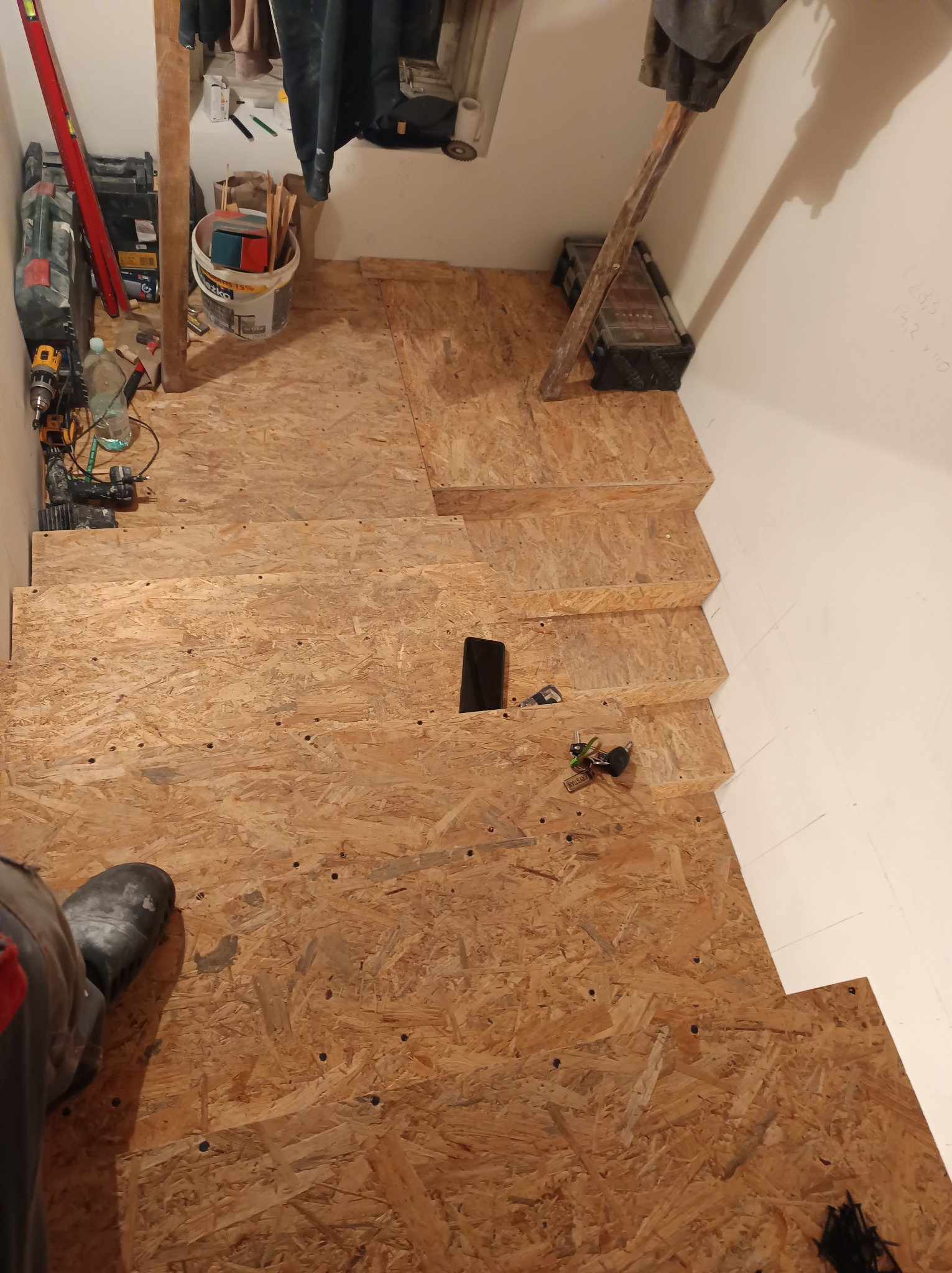 Szkielet schodów z płyty OSB w trakcie budowy, widoczne narzędzia, wiertarka, puszka z farbą i częściowo widoczna noga w bucie roboczym.