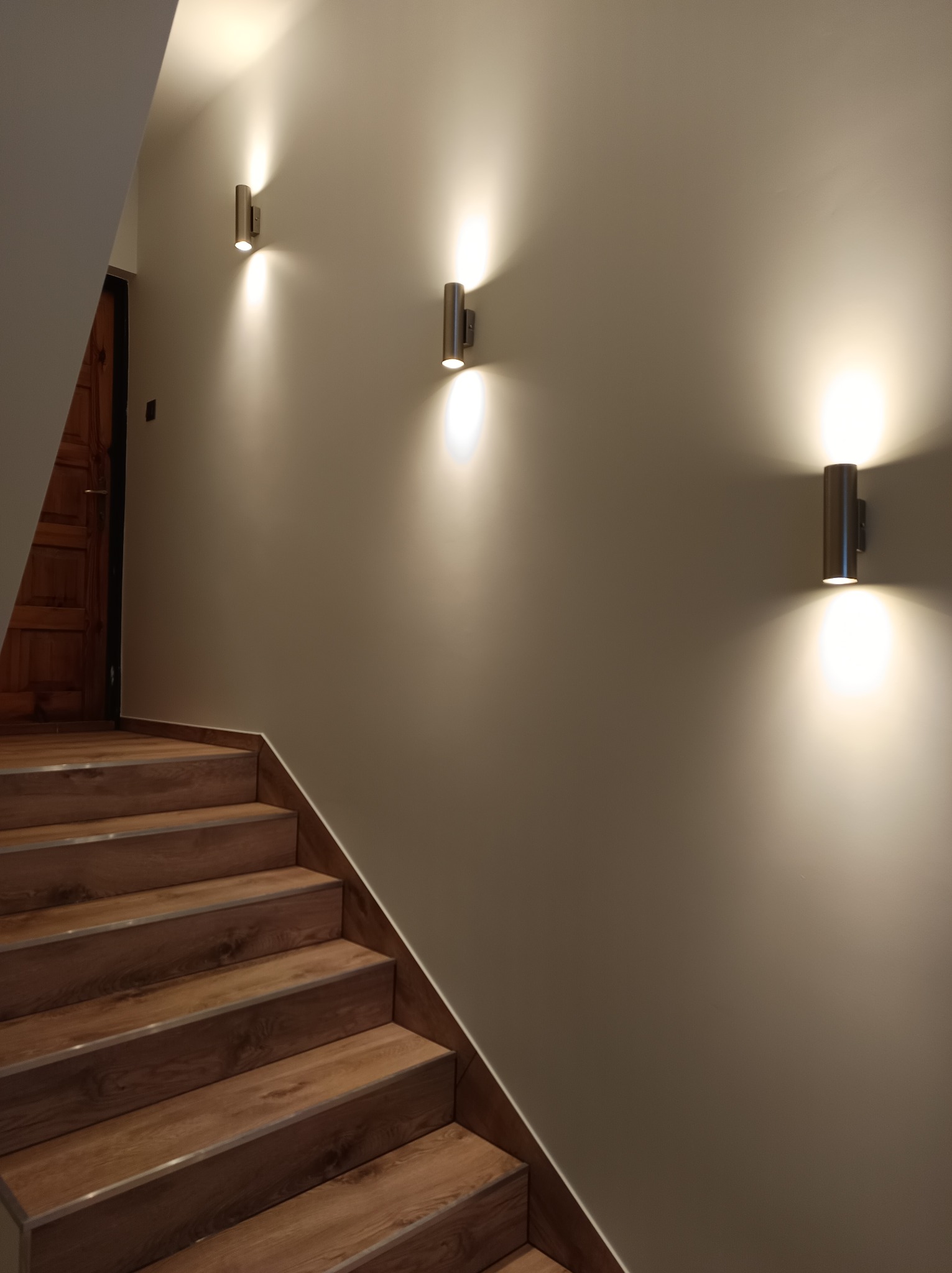 Klatka schodowa z drewnopodobnymi schodami, oświetlona trzema cylindrycznymi kinkietami emitującymi światło w górę i w dół, ściana pomalowana na jasny beż.