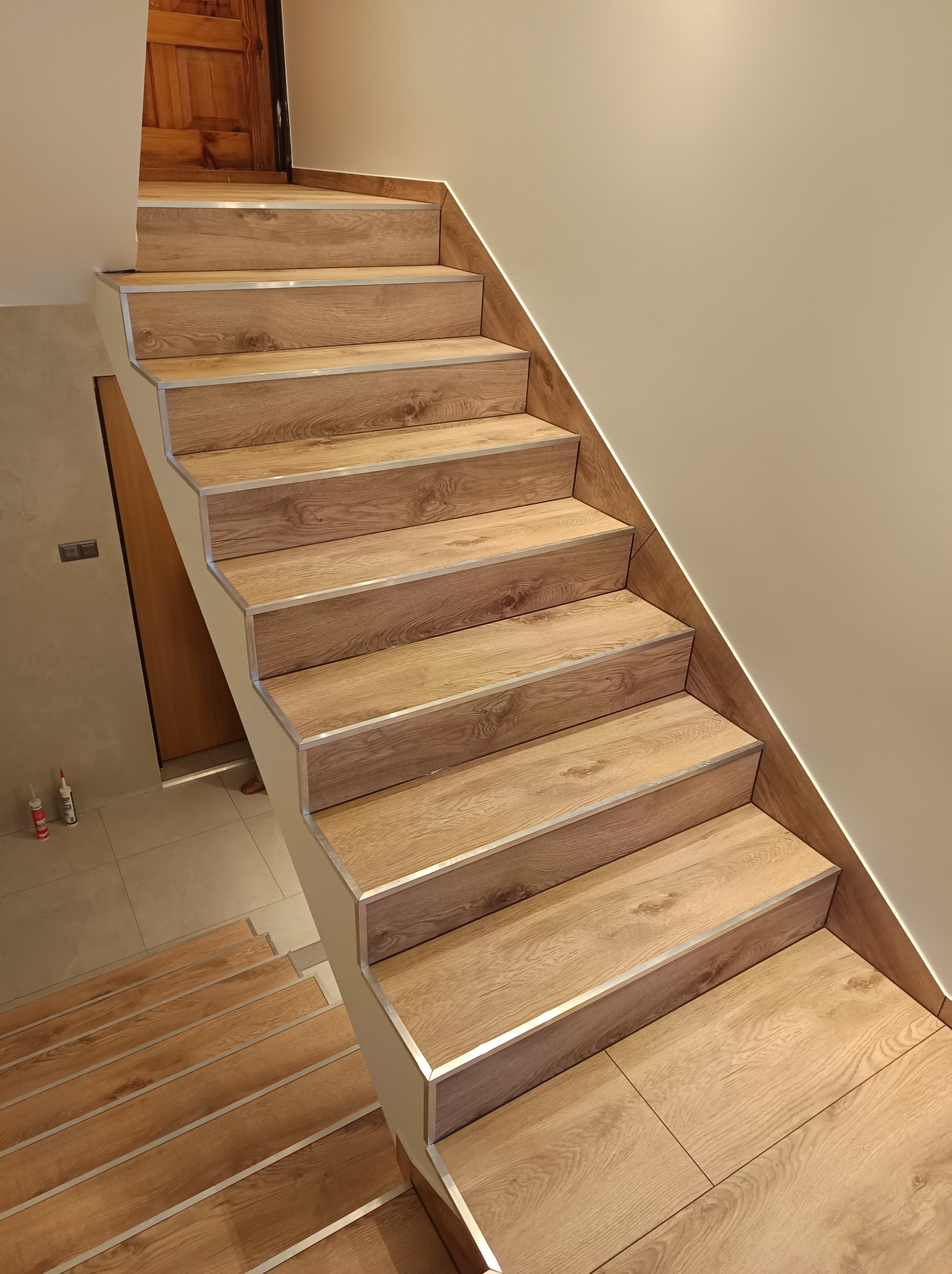 Schody wykończone panelami imitującymi drewno, z aluminiowymi listwami na krawędziach stopni, oświetlenie LED wzdłuż ściany, widok z dołu na górę.