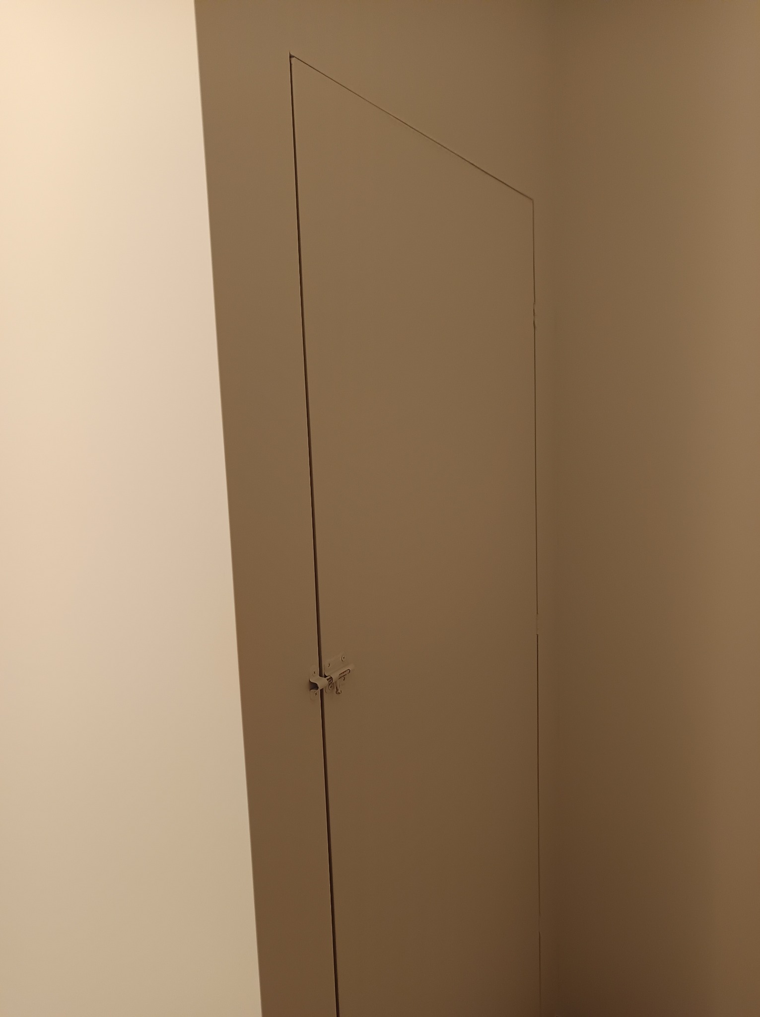 Ukryte drzwi do schowka na poddaszu z zamkiem baskwilowym, wtopione w ścianę pomalowaną na jednolity kolor.