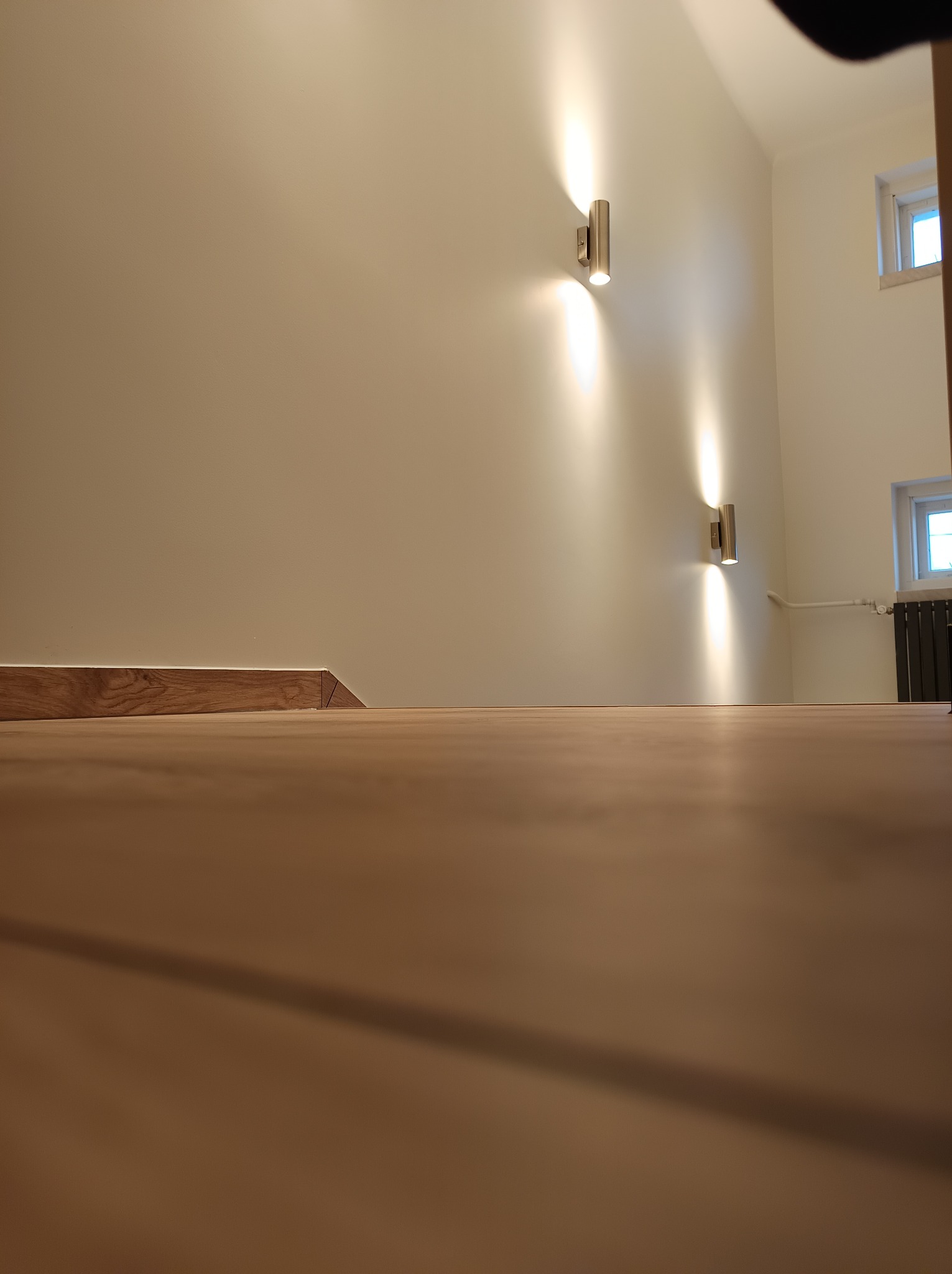 Widok z poziomu podłogi na świeżo pomalowaną ścianę z dwoma nowoczesnymi, metalowymi kinkietami, drewnianą listwą przypodłogową i grzejnikiem pod oknem.