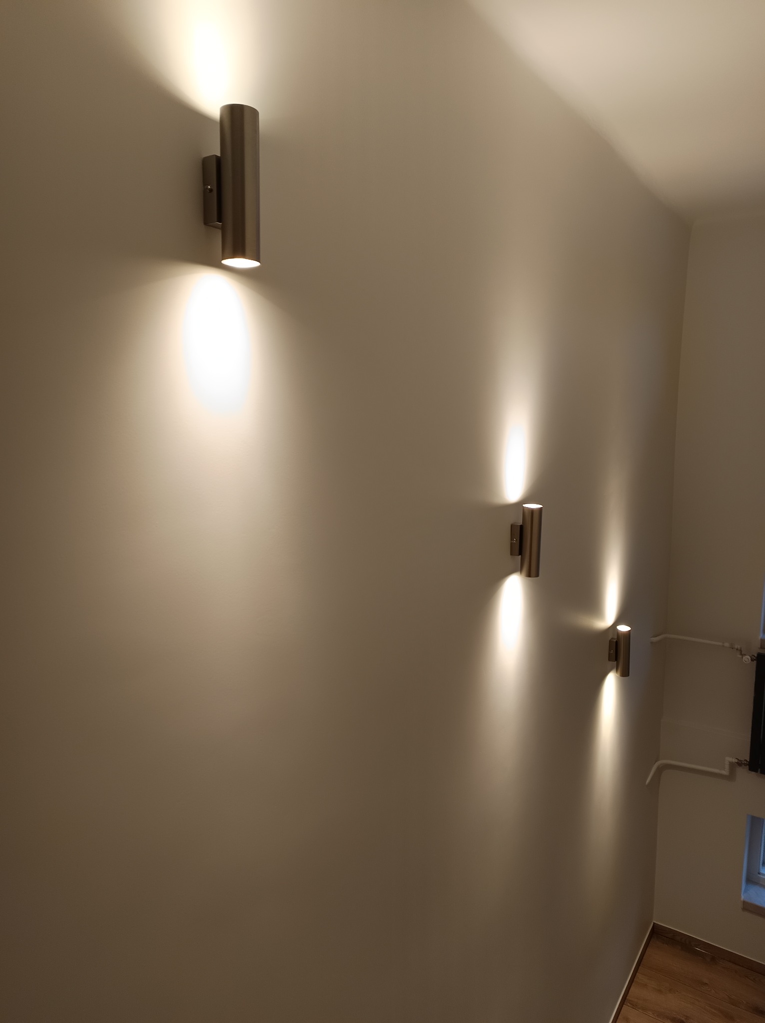 Ściana z trzema metalowymi lampami ściennymi o cylindrycznym kształcie, emitującymi ciepłe światło w górę i w dół, oświetlającymi białą ścianę.