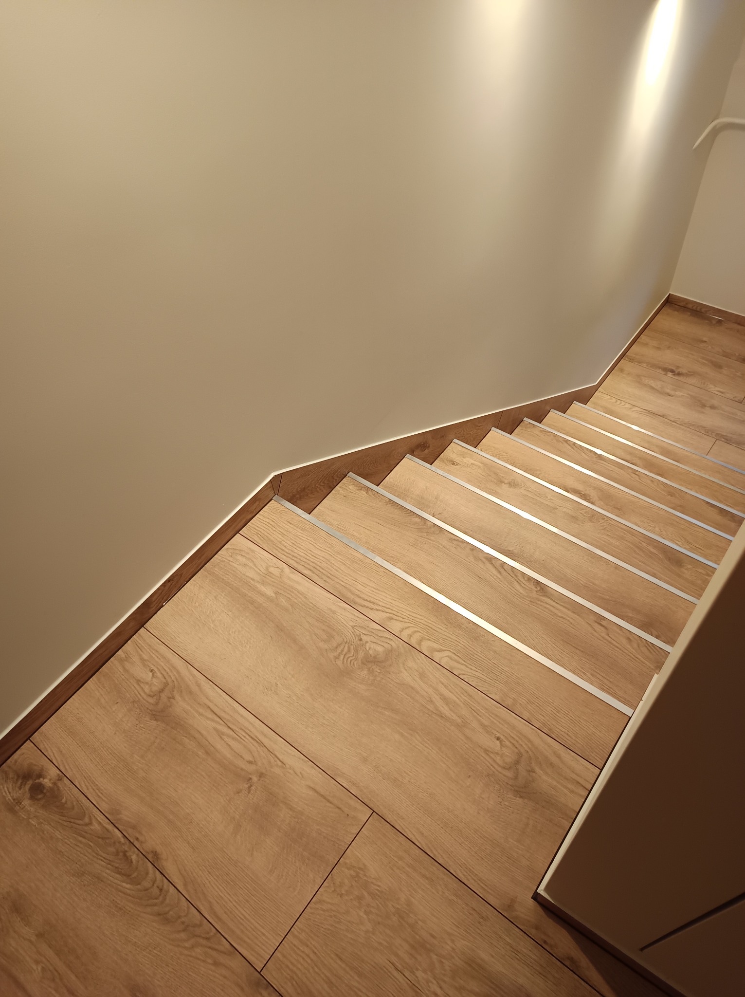 Schody wykończone panelami drewnopodobnymi z metalowymi listwami antypoślizgowymi, widok z góry, oświetlenie punktowe na ścianie.