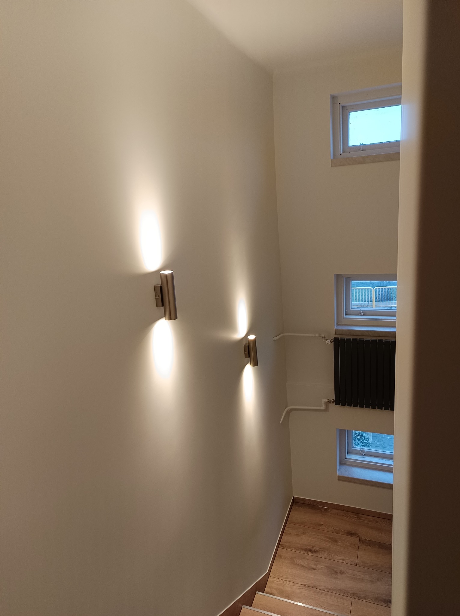 Klatka schodowa z oświetleniem ściennym w postaci dwóch metalowych lamp cylindrycznych, trzema małymi oknami i czarnym grzejnikiem żeberkowym pod jednym z nich, wykończona w jasnych odcieniach.