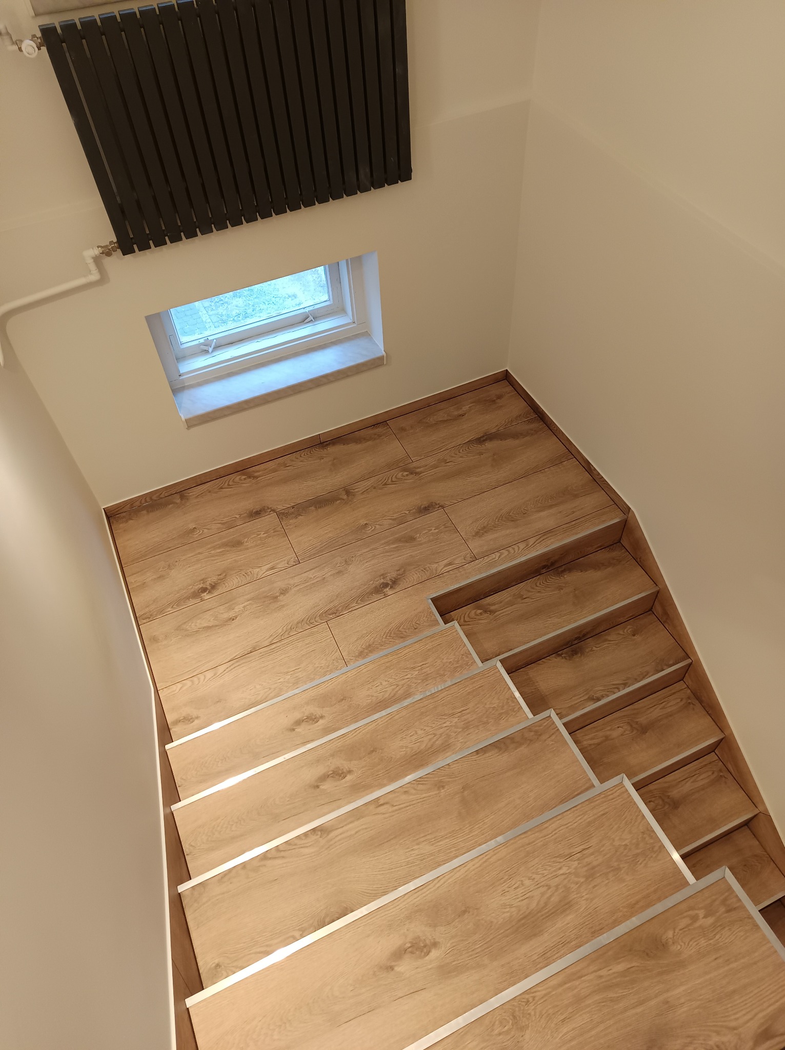 Wykończone schody wewnętrzne z drewnopodobnymi panelami, zabezpieczone srebrnymi listwami, widok z góry, na ścianie grzejnik i małe okno.