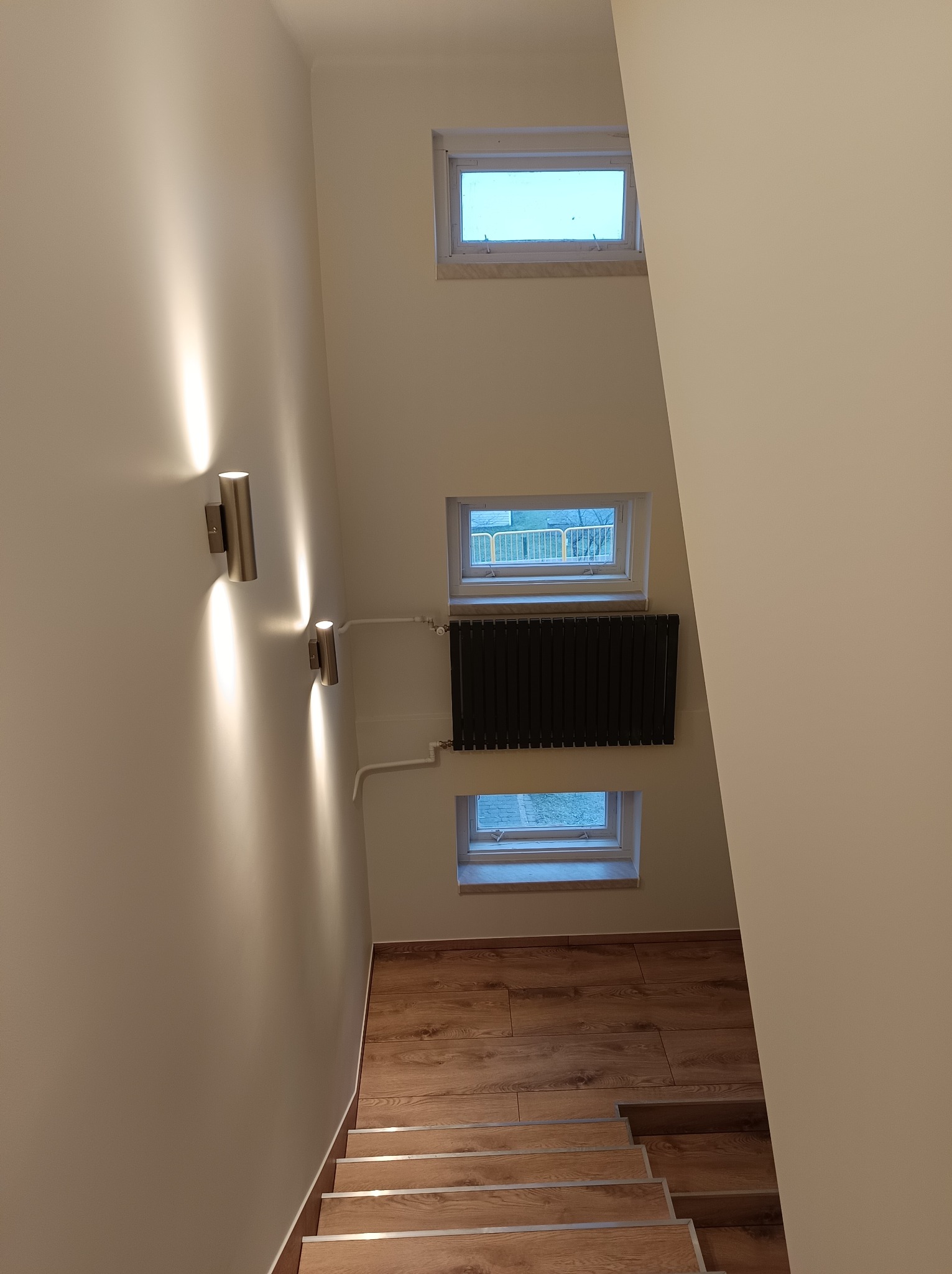 Klatka schodowa z drewnianymi schodami i trzema małymi oknami, oświetlona dwoma nowoczesnymi kinkietami.