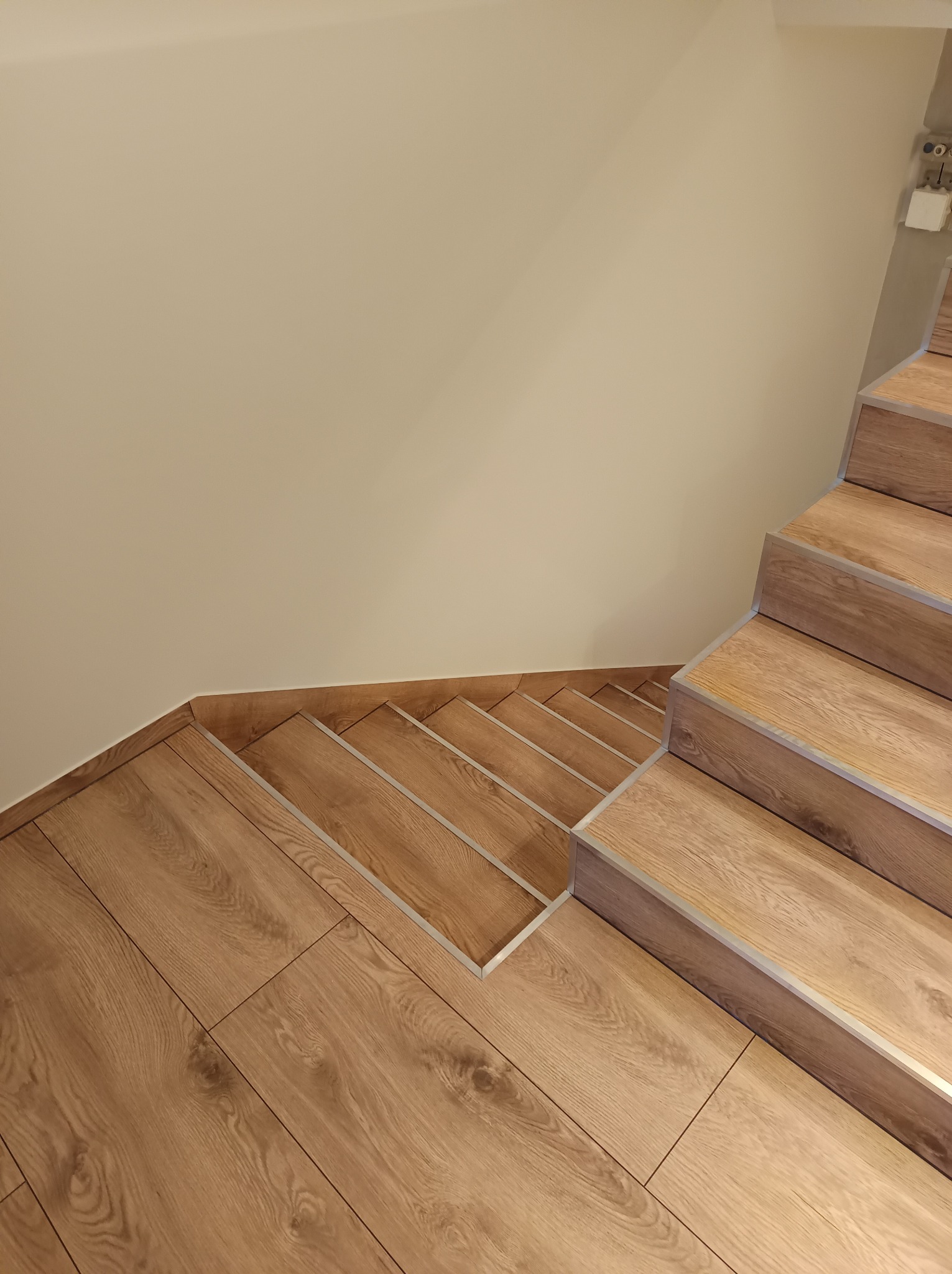 Wykończone schody drewnopodobne z listwami aluminiowymi, prowadzące w dół, z widoczną podłogą w tym samym stylu i kremową ścianą w tle.