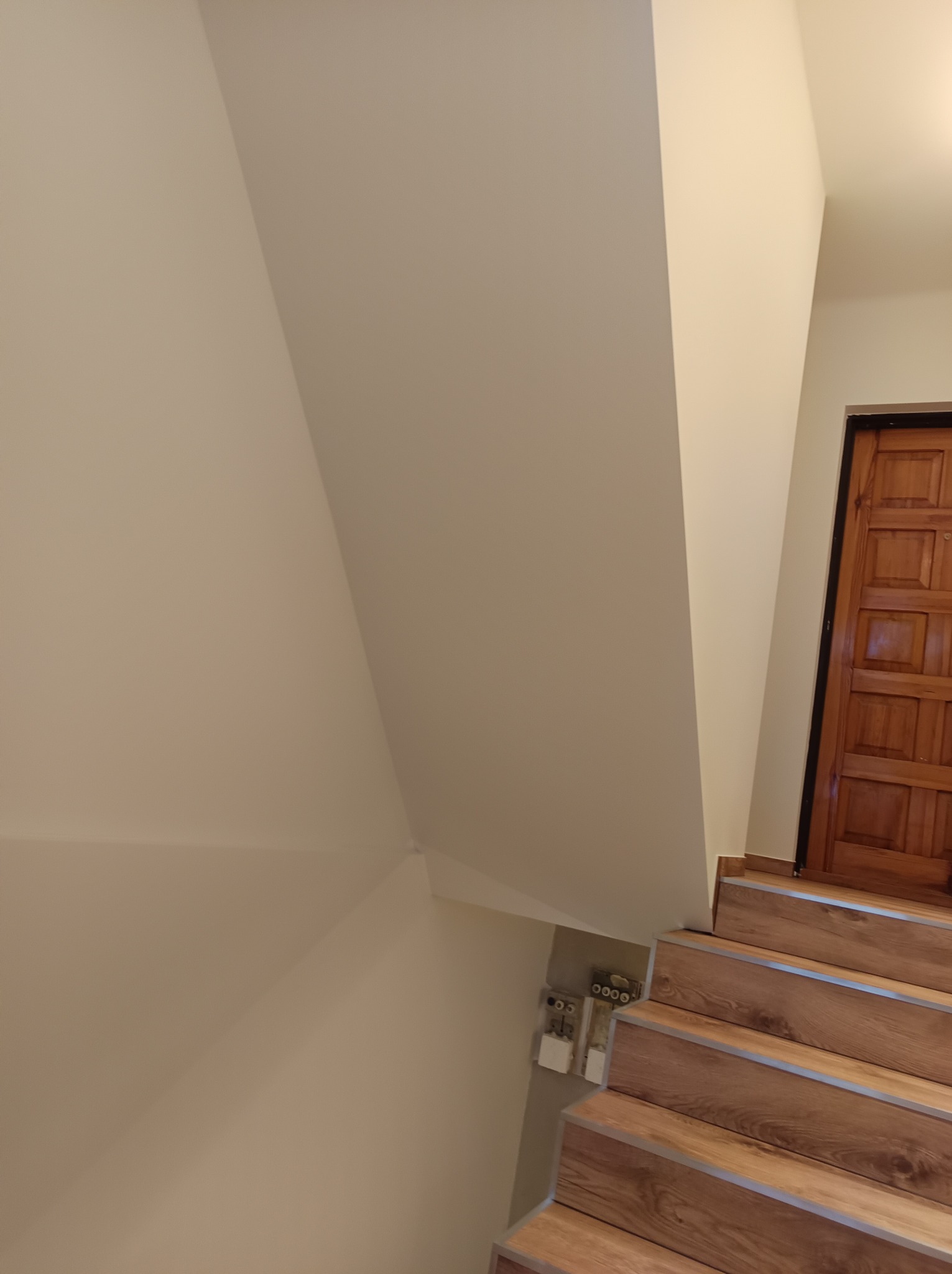 Klatka schodowa po remoncie z drewnopodobnymi panelami na schodach i świeżo pomalowanymi ścianami w odcieniach beżu, widoczne gniazdka elektryczne. Nowoczesne wykończenie.