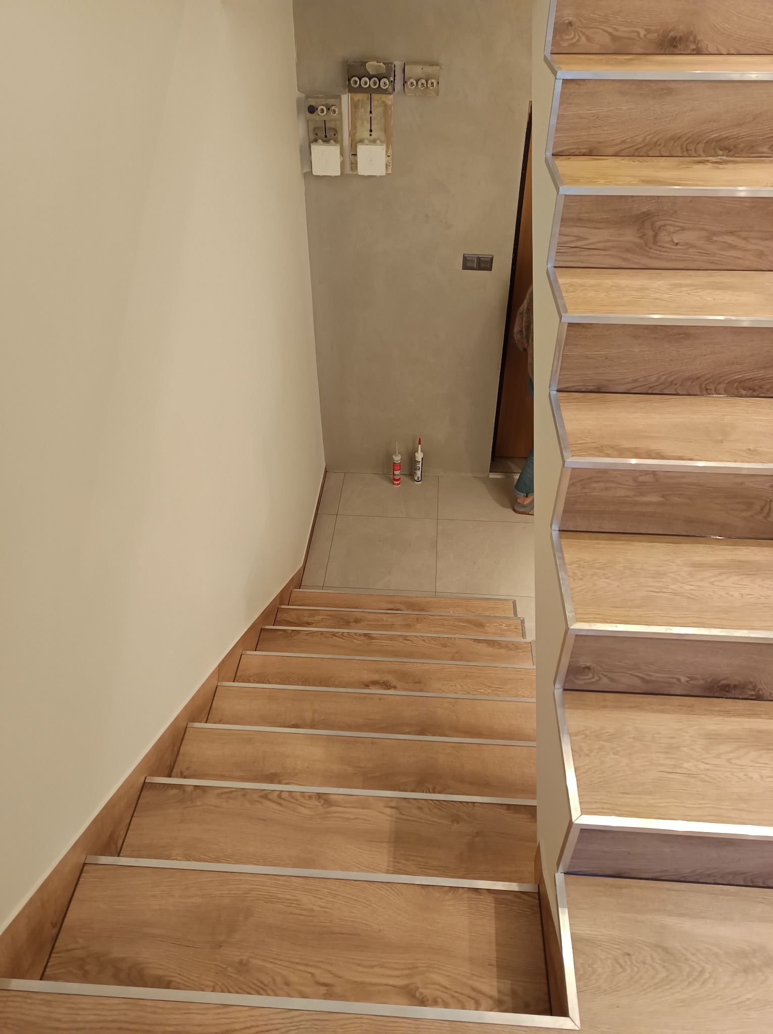 Wykończone schody drewniane z metalowymi listwami zabezpieczającymi krawędzie, widok z góry, na ścianie widoczne puszki elektryczne i dwie tuby kleju silikonowego.