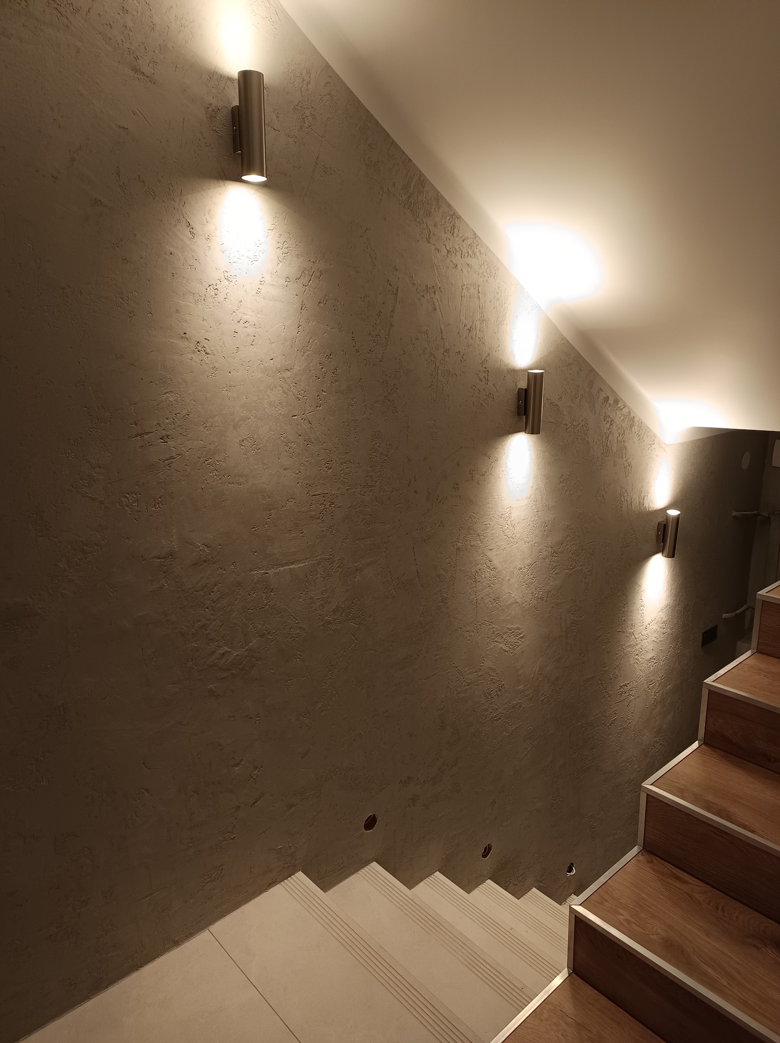 Ściana z dekoracyjną strukturą oświetlona trzema podłużnymi lampami ściennymi na klatce schodowej w Zbijowie Małym. Widoczne drewniane stopnie schodów.