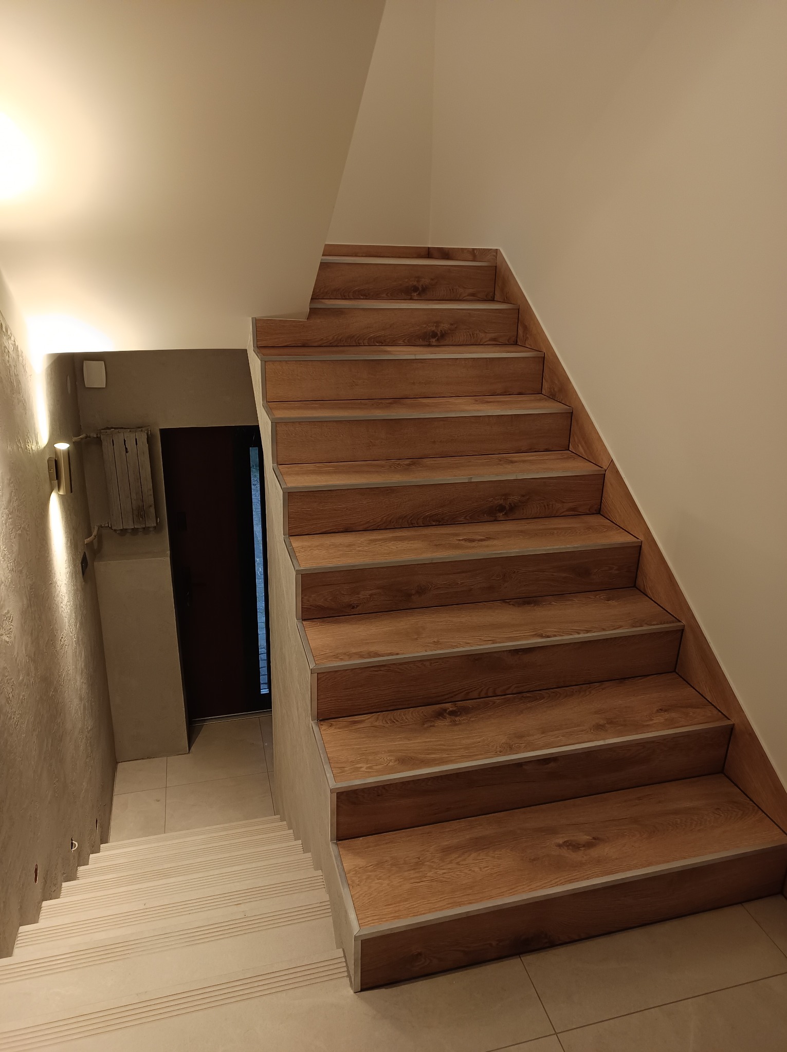 Schody wewnętrzne z drewnopodobnymi panelami, widok z góry, z delikatnym oświetleniem punktowym na ścianie obok, kontrastujące z białymi stopniami dolnymi i jasnymi ścianami.