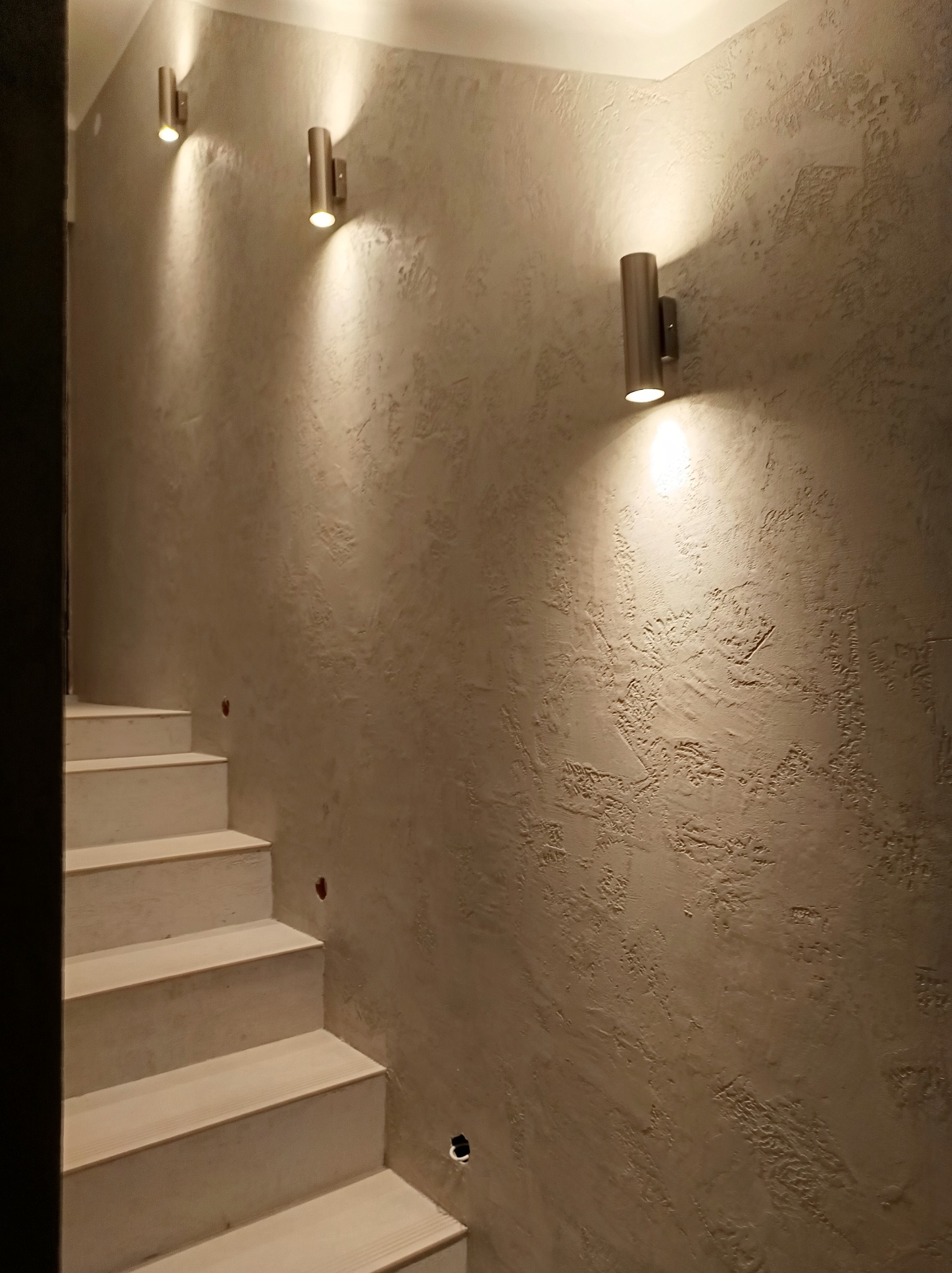 Ściana z dekoracyjną fakturą i trzema metalowymi kinkietami o cylindrycznym kształcie, oświetlającymi schody z jasnego drewna.