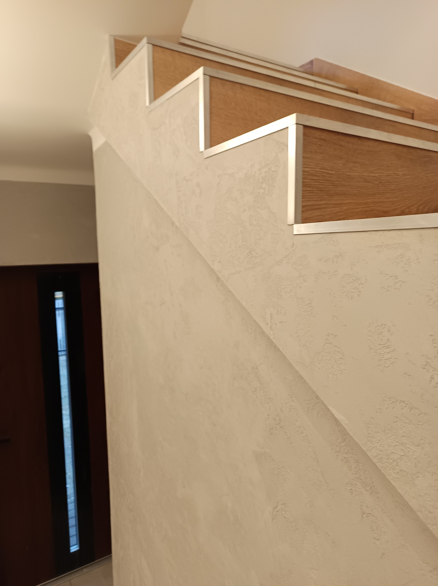 Schody wewnętrzne z drewnianymi stopniami i metalowym wykończeniem, ściana z dekoracyjną fakturą w odcieniach beżu, widoczny fragment drzwi.