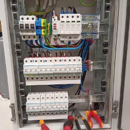 Otwarta skrzynka elektryczna z zainstalowanymi bezpiecznikami, ogranicznikami przepięć i okablowaniem, widoczne narzędzia: kombinerki i śrubokręty.