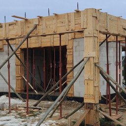 Construction of the house - Budowa domu: szalunki z drewna wsparte na podporach, białe bloczki fundamentowe, widoczna osoba w ciemnym ubraniu. Zima, ziemia pokryta śniegiem.