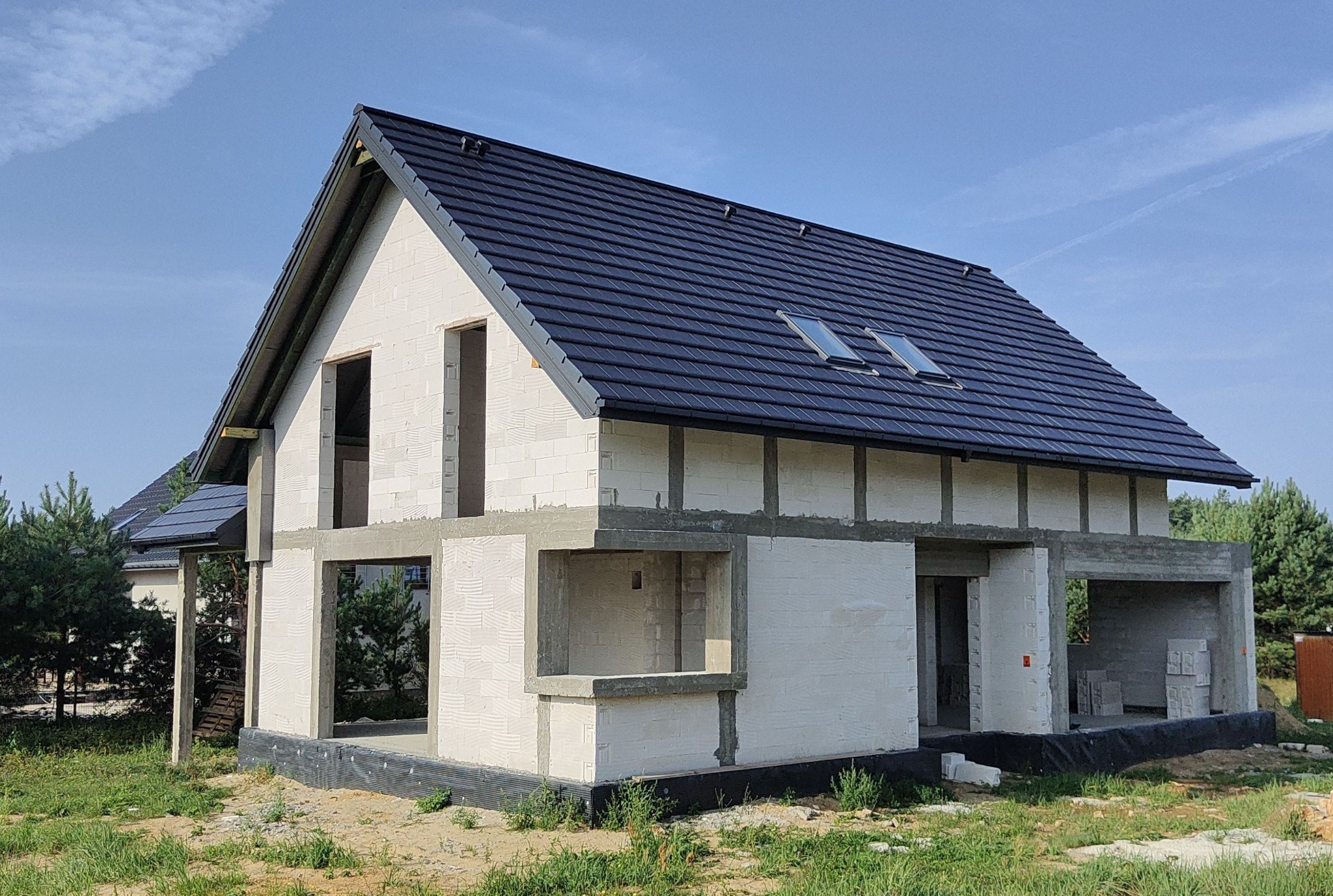 Surowy, dwukondygnacyjny dom jednorodzinny w trakcie budowy, z ciemną dachówką i oknami dachowymi, wzniesiony z białych bloczków, widoczne niezadaszone otwory okienne i drzwiowe, na tle błękitnego...