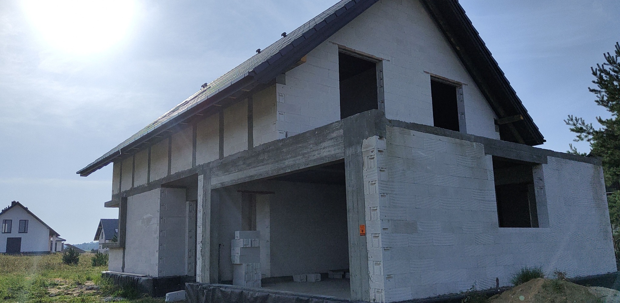 Surowy stan domu murowanego z betonu komórkowego, widoczne otwory okienne i brakujące elementy elewacji, w tle inne domy w zabudowie jednorodzinnej.
