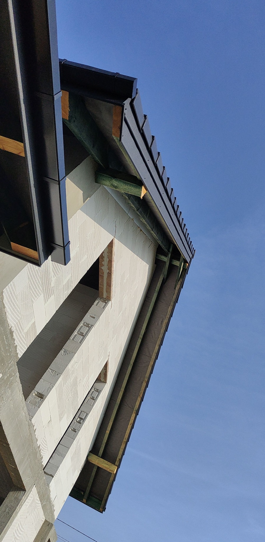 Surowy stan domu z białych bloczków, widoczne okna bez ram, czarne rynny i krokwie na tle błękitnego nieba.