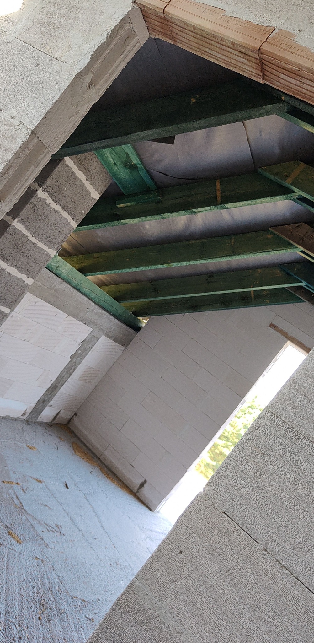 Wnętrze poddasza w trakcie budowy: widoczne krokwie pomalowane na zielono, ściany z białych bloczków i fragmenty muru z szarych pustaków.