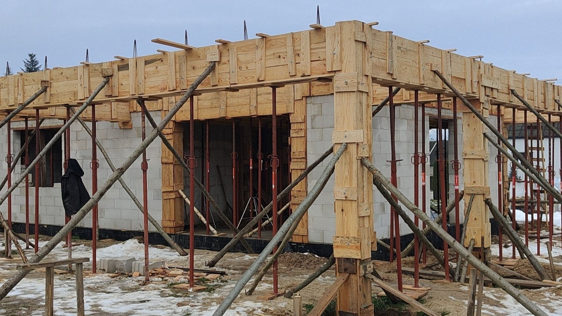 Budowa domu: szalunki z drewna wsparte na podporach, białe bloczki fundamentowe, widoczna osoba w ciemnym ubraniu. Zima, ziemia pokryta śniegiem.